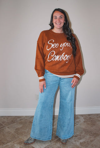 Person wearing a brown sweatshirt with 'See you Cowboy!' text and blue jeans against a plain background